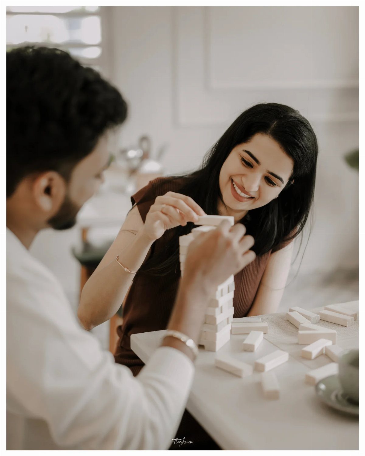 Couple smiling and playing Jenga with wooden blocks on a white table indoors.