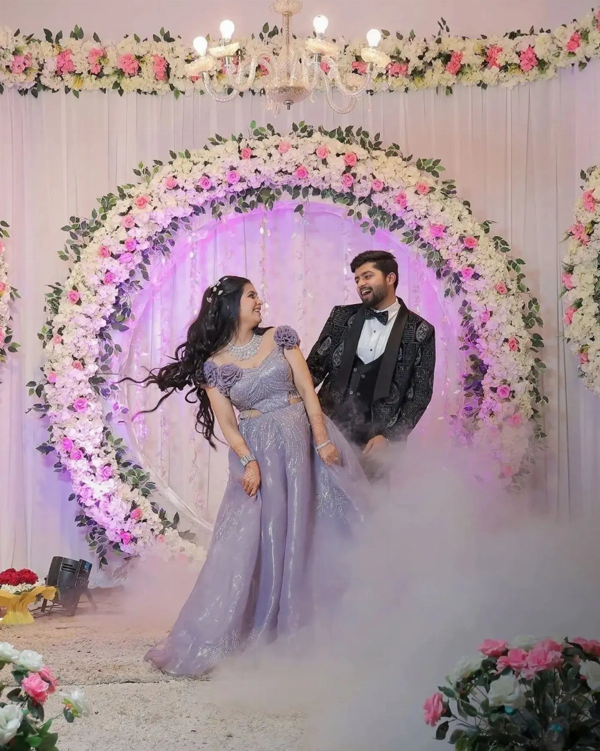 Couple posing in formal attire before a floral arch with white floor fog.