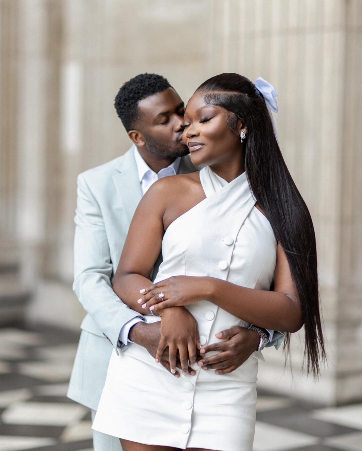 Man in light blue suit embracing woman in white dress and kissing her cheek.