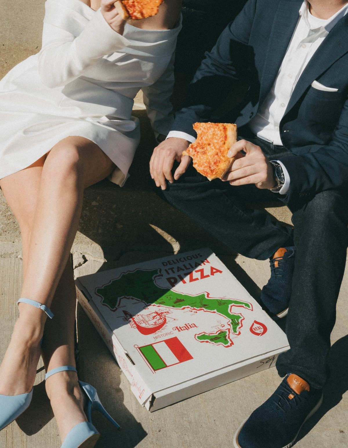 Couple in formal attire sitting on concrete eating pizza from a cardboard box
