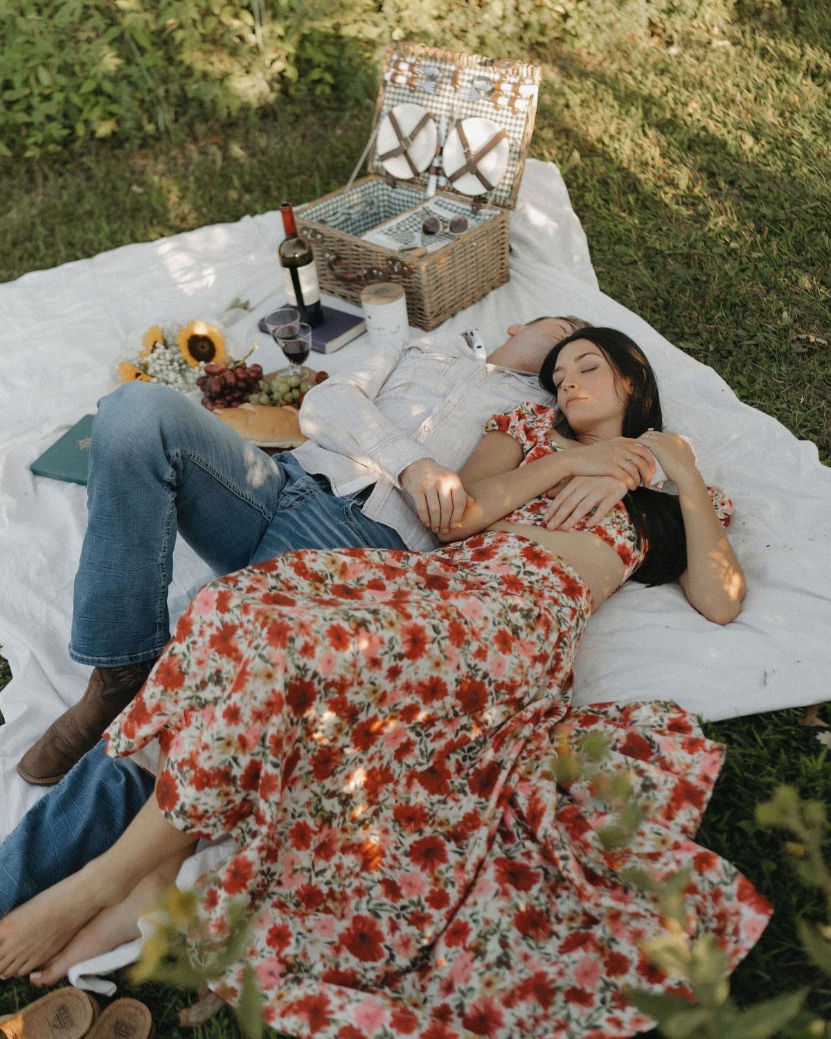 Couple lying side by side on a picnic blanket in a sunny grassy field.