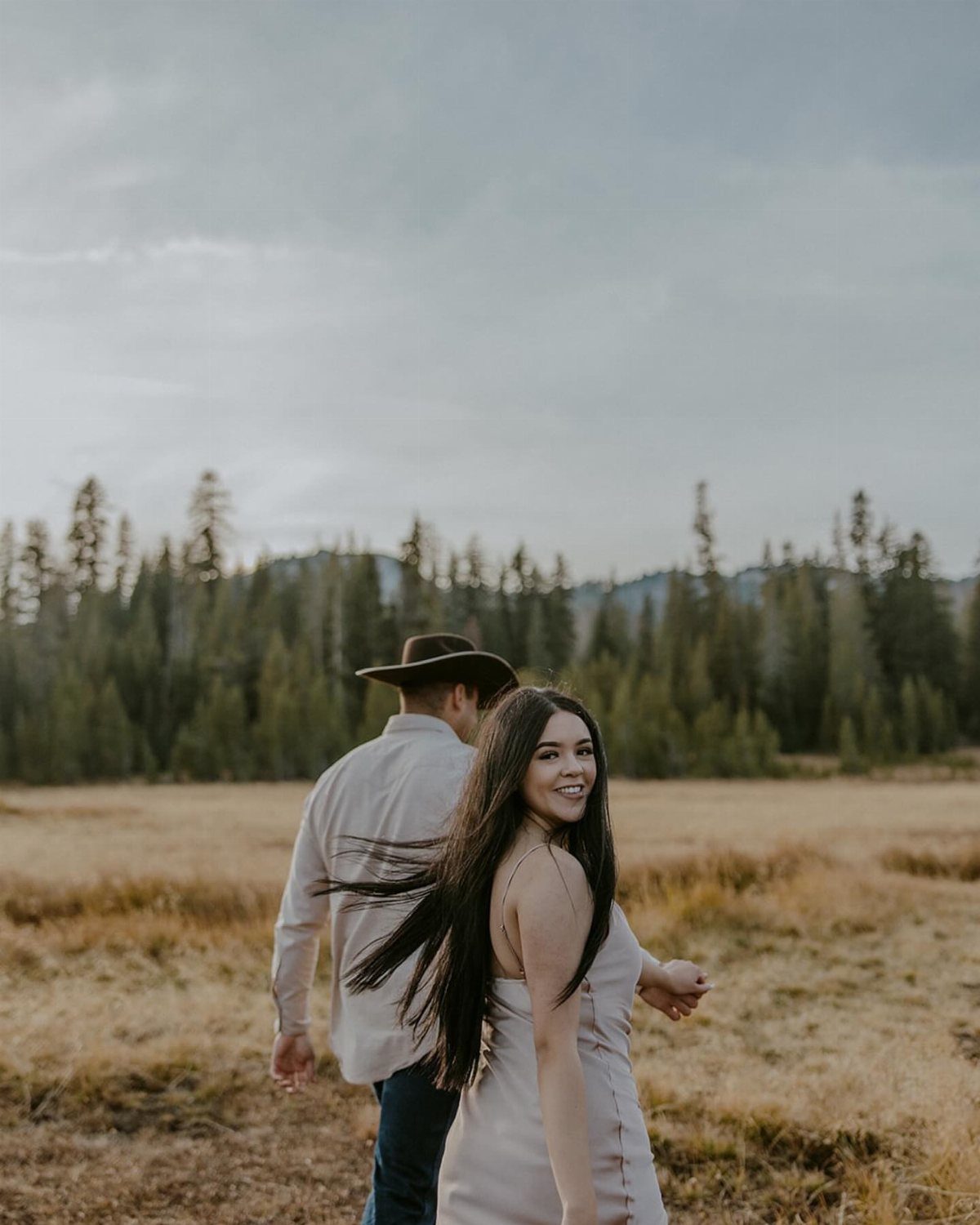 Couple walking through a golden field with the woman looking back and smiling.