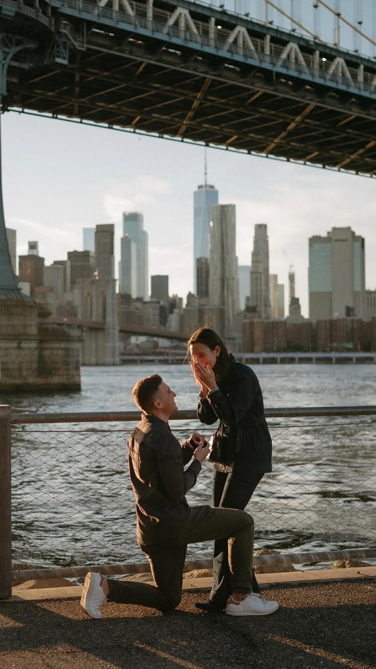 Man proposing to woman on a waterfront with the Manhattan skyline and bridge.