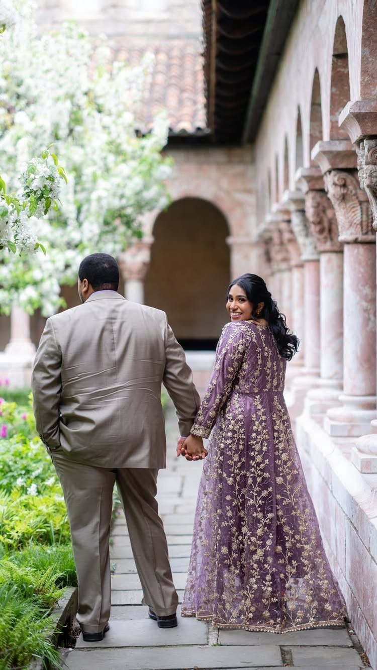 Couple in formal attire holding hands in a stone courtyard with arched columns.