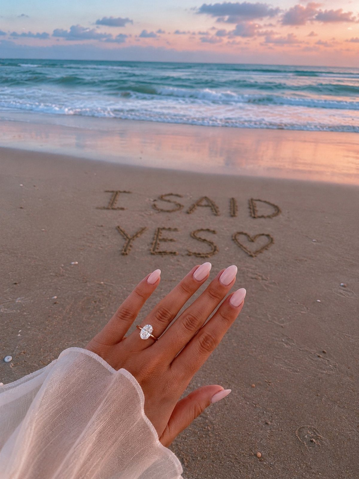 Hand with diamond engagement ring over 'I SAID YES' written in beach sand.