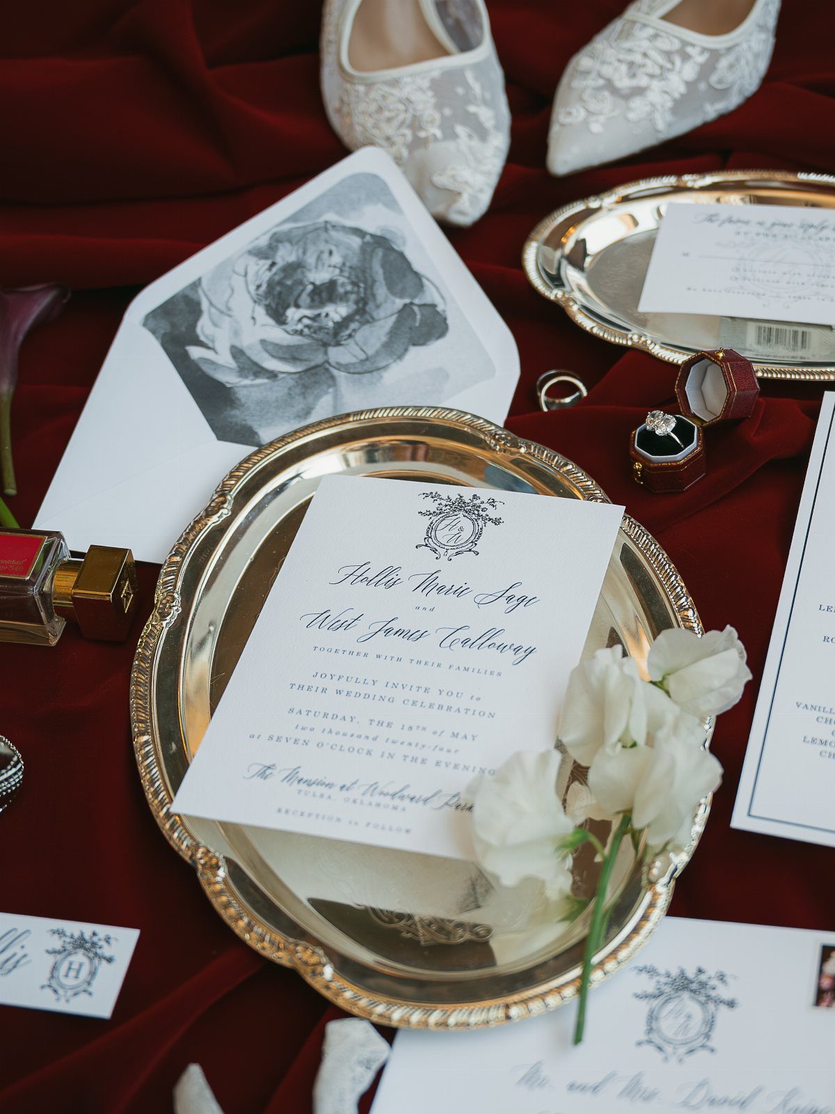Wedding flat lay with engagement ring, lace shoes, and invitations on red fabric.