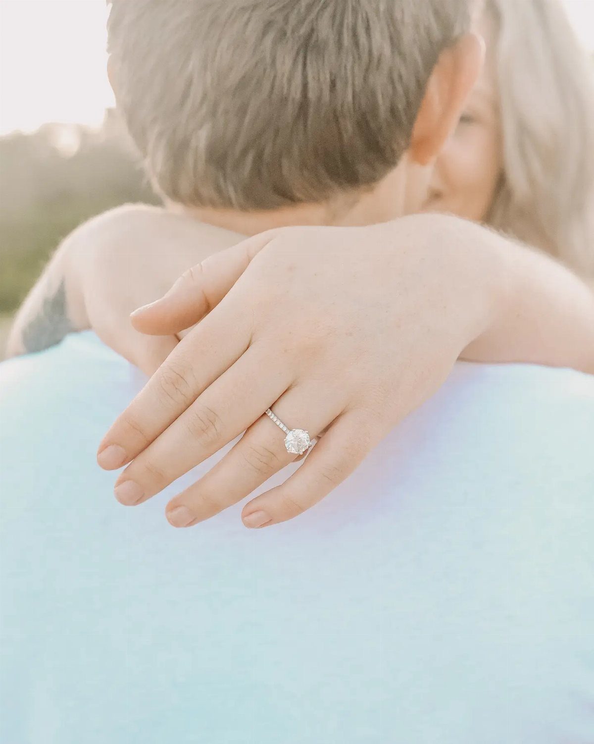 Hand with diamond engagement ring resting on a partner's light blue shoulder