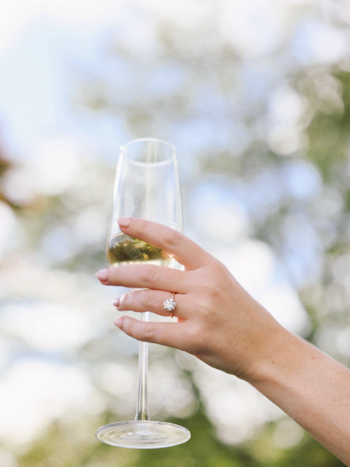 Hand holding a champagne flute with a diamond engagement ring on the finger