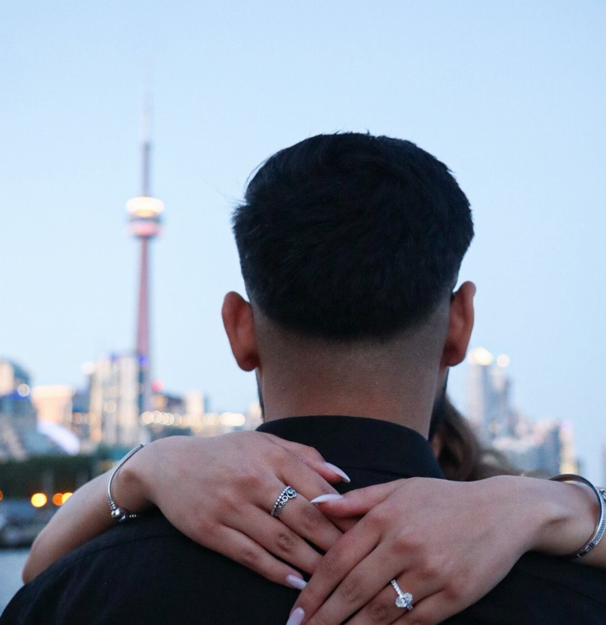 Couple hugging from behind with a diamond ring visible against a city skyline.