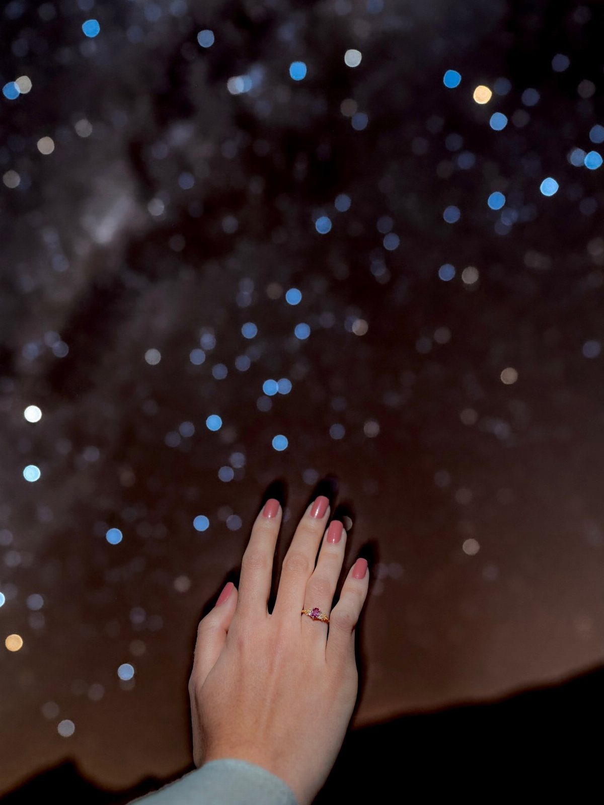 Hand wearing a gold ring with purple stone against a starry night sky.