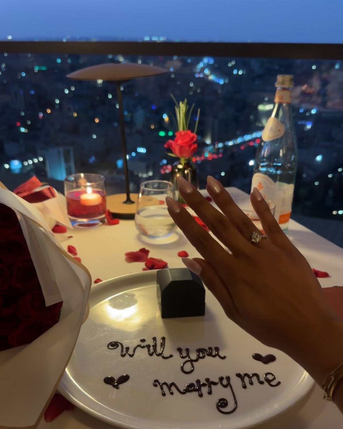 Hand with engagement ring over a proposal plate with a city skyline background