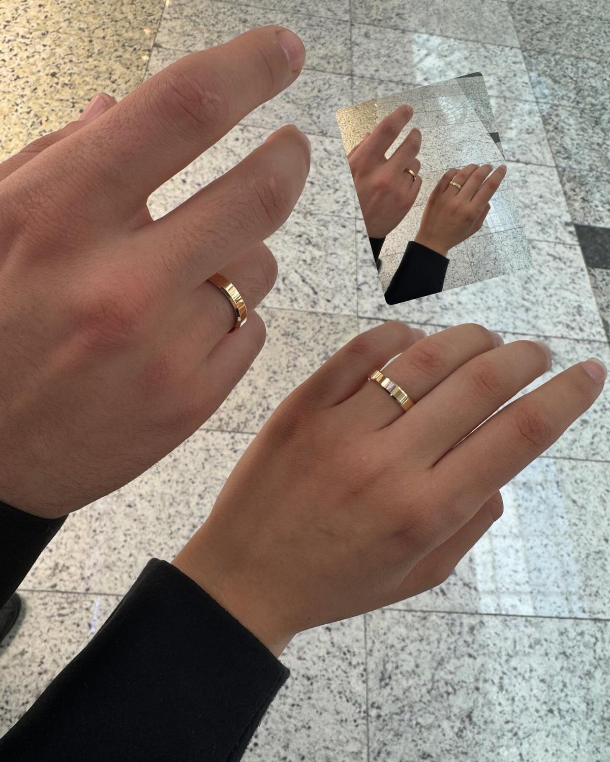 Two hands with gold rings reflected in a small mirror on tiled flooring