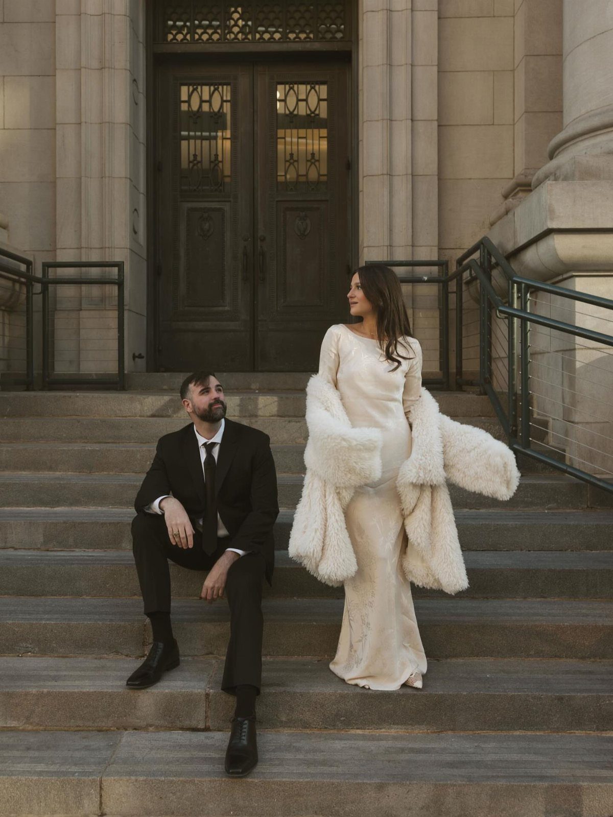 Couple posing on stone steps, woman wearing a white dress and faux-fur coat.