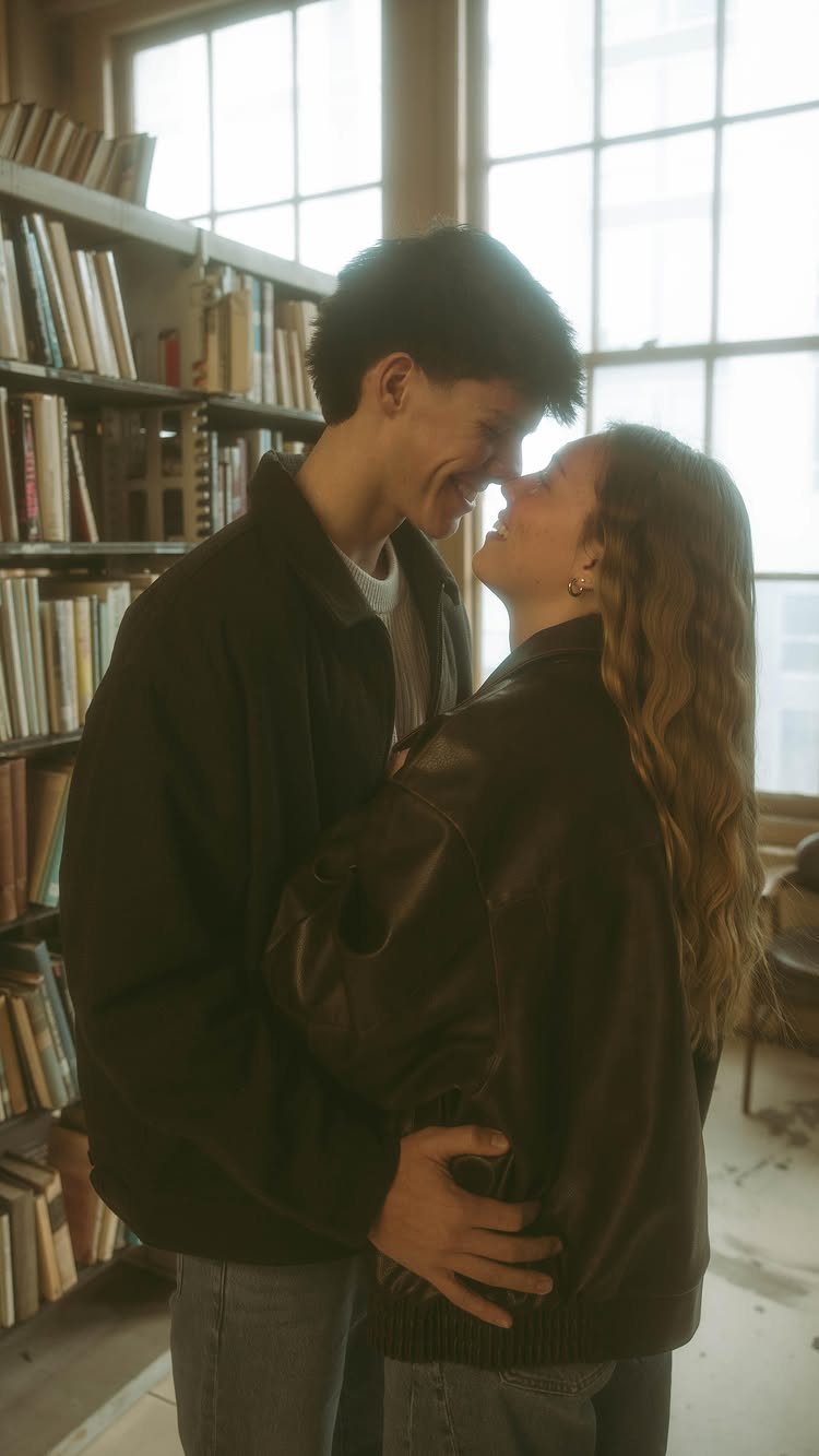 Young couple embracing and smiling in a cozy library with bookshelves and windows.