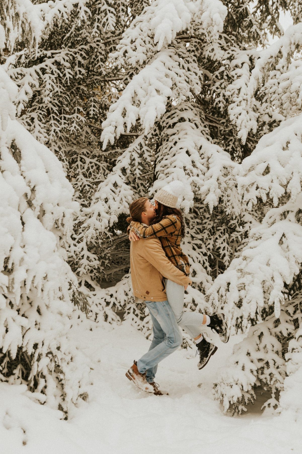 Couple kissing in a snowy forest with snow-covered evergreen trees surrounding them.
