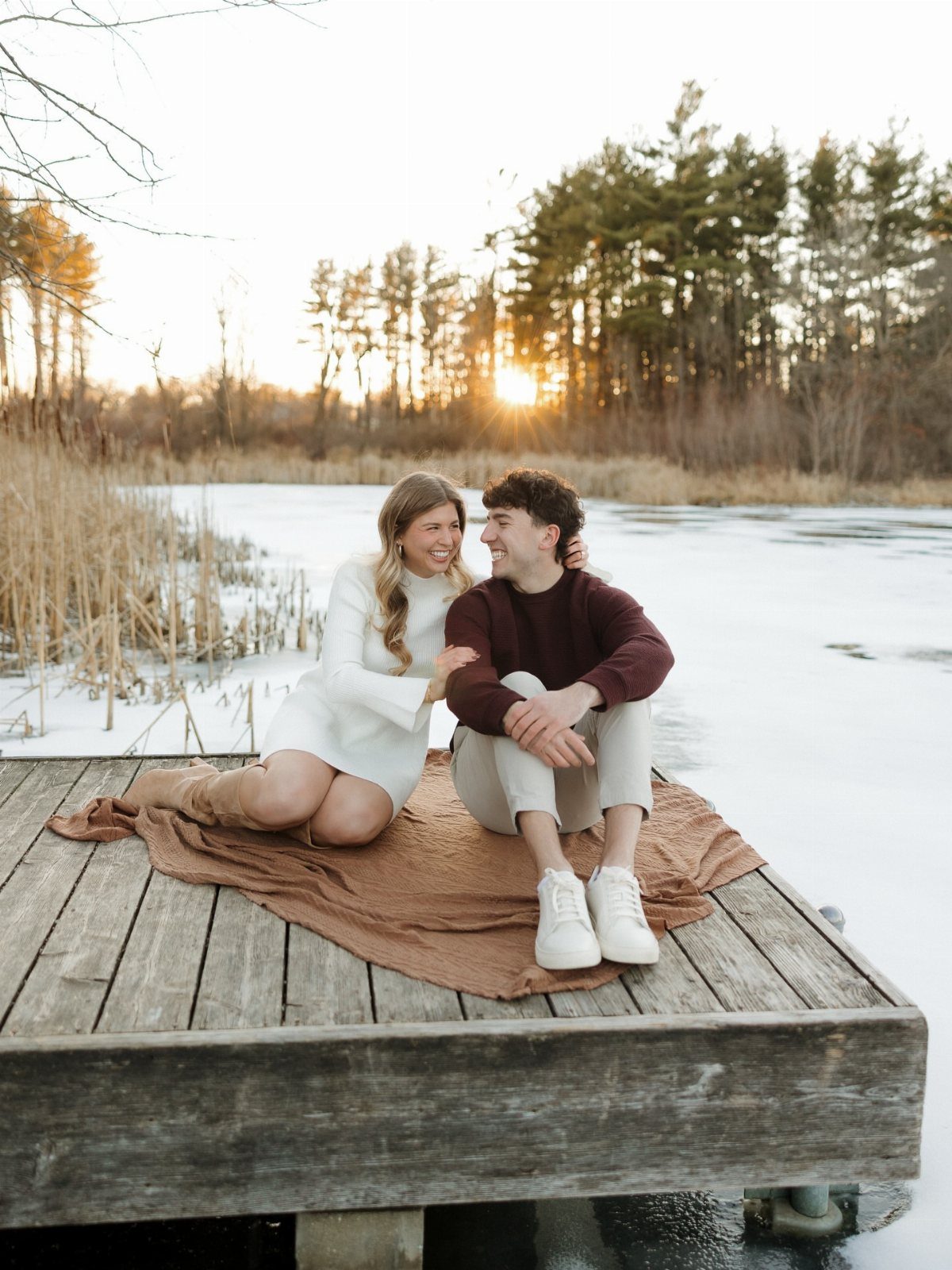 Smiling couple sitting on a blanket on a wooden dock by a frozen lake.