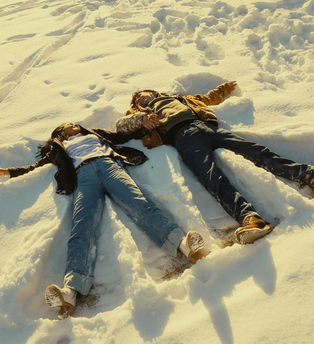 Couple lying side-by-side in deep white snow wearing winter jackets and jeans.