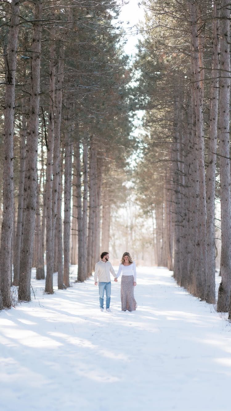 Couple holding hands walking through a snowy forest path lined with tall pine trees
