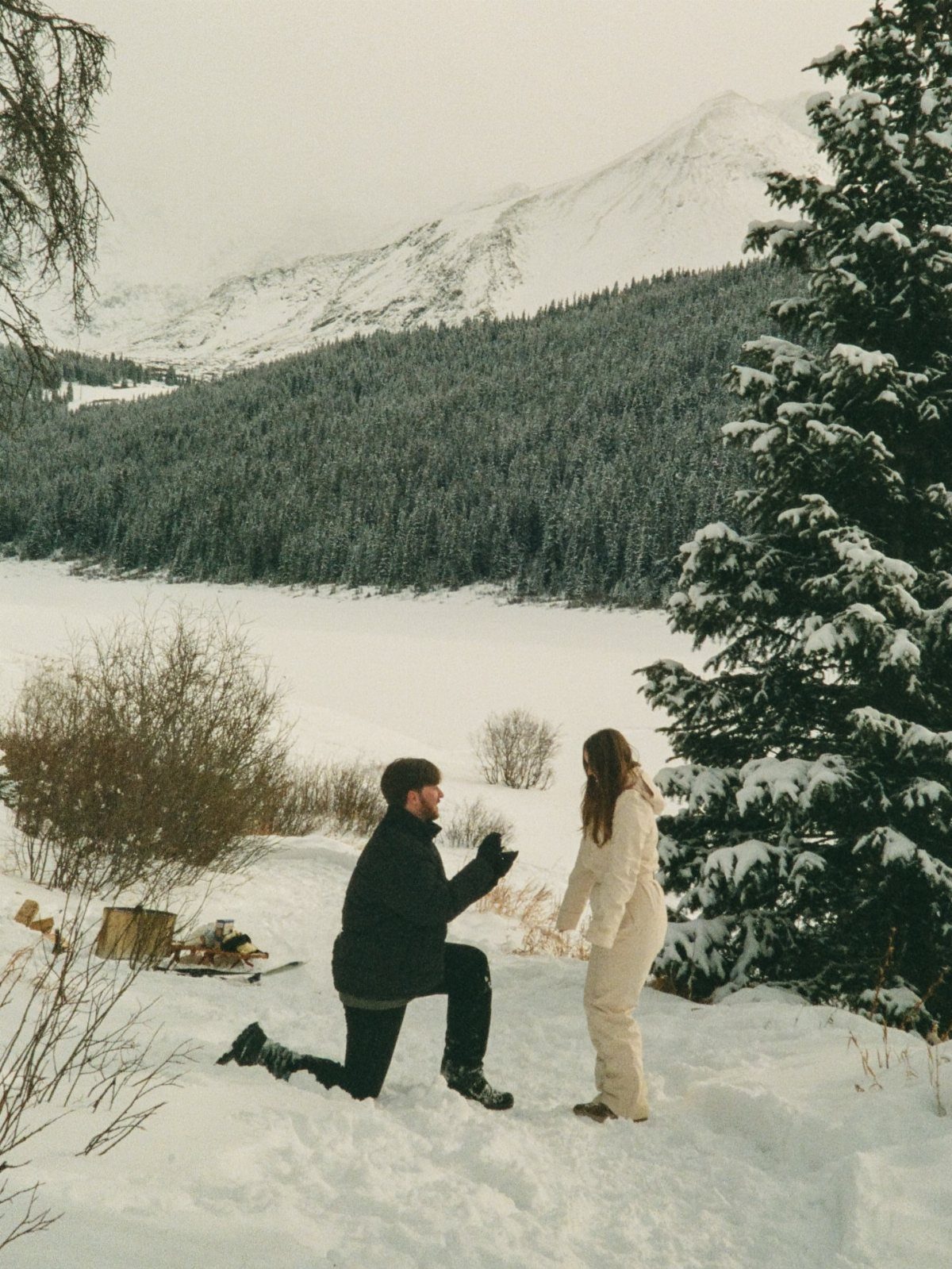 Man proposing to a woman in a snowy mountain landscape with evergreen trees.