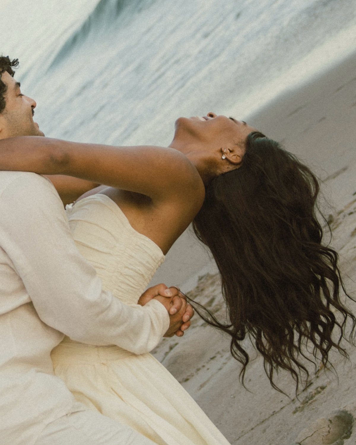 Couple in white clothing embracing on a beach with the woman leaning back.