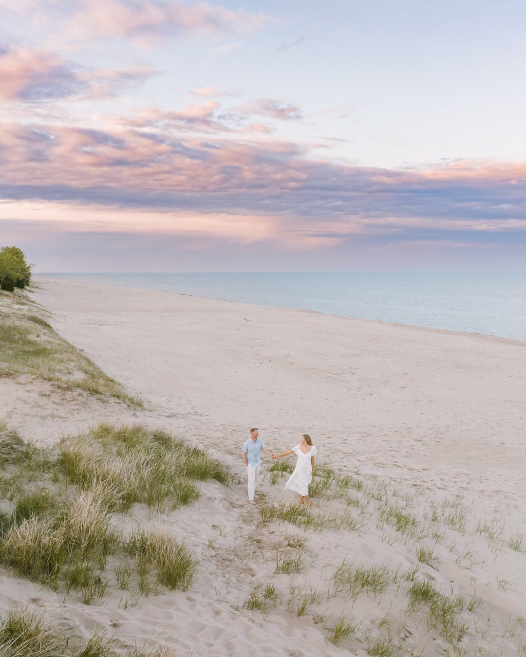 Couple holding hands on a sandy beach with dunes under a sunset sky