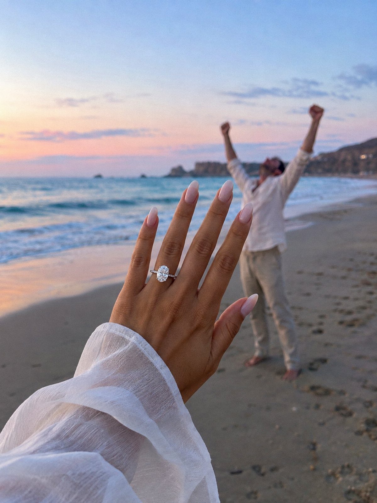 Woman's hand with engagement ring in foreground, man cheering on beach at sunset