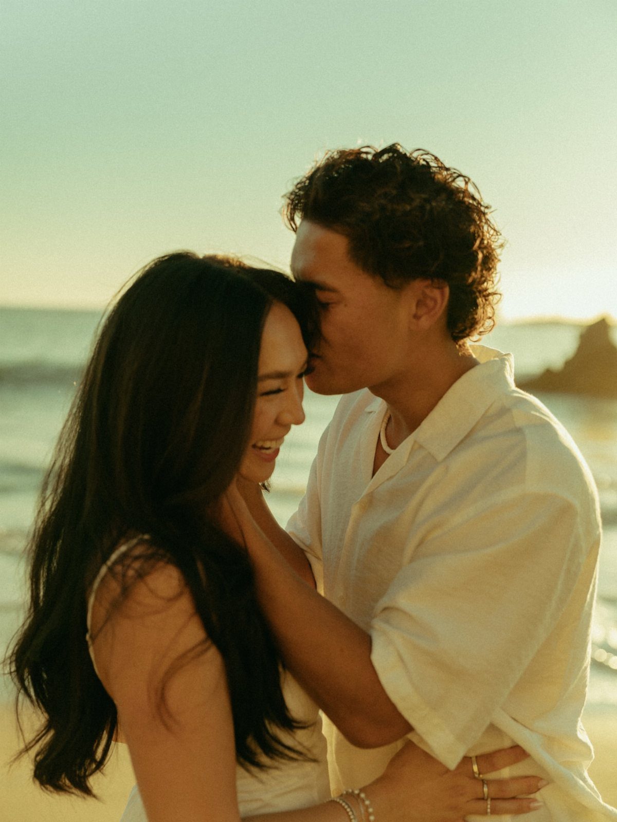 Couple embracing on a beach at sunset, man kissing woman's forehead.