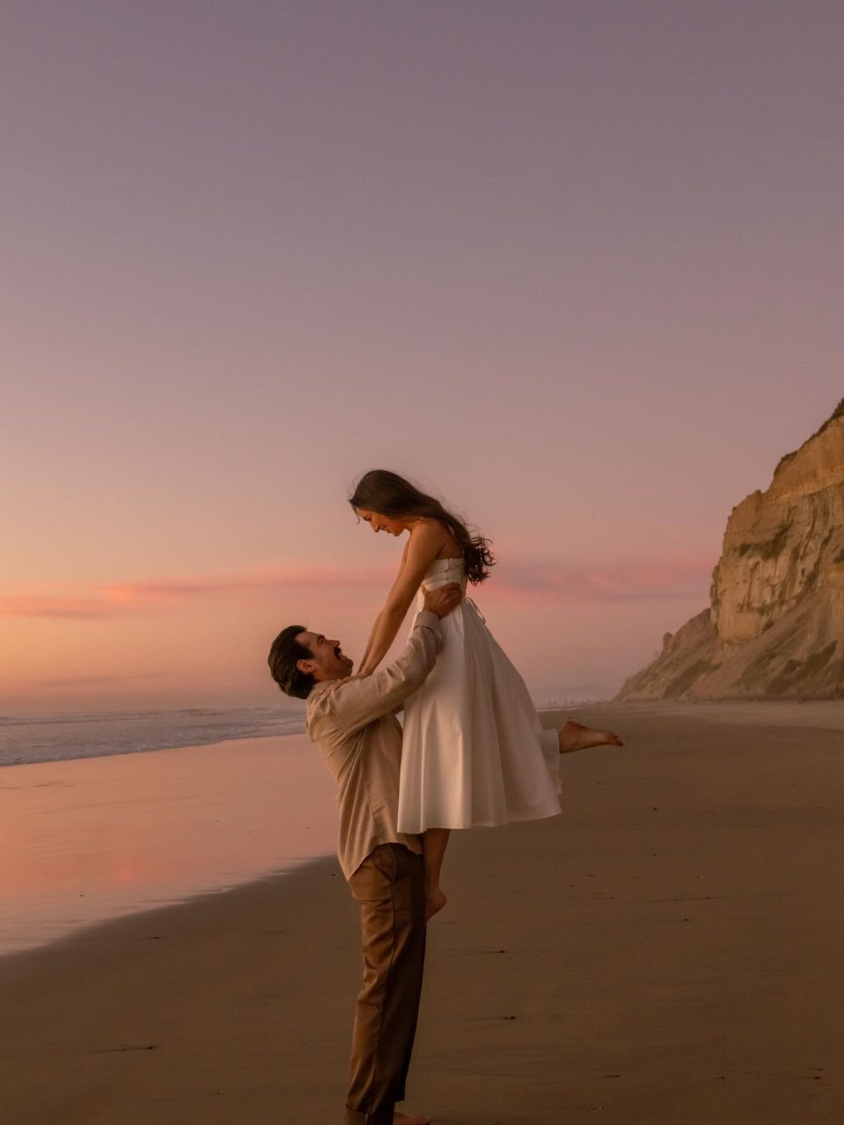 Man lifting woman in white dress on a beach during a sunset photoshoot