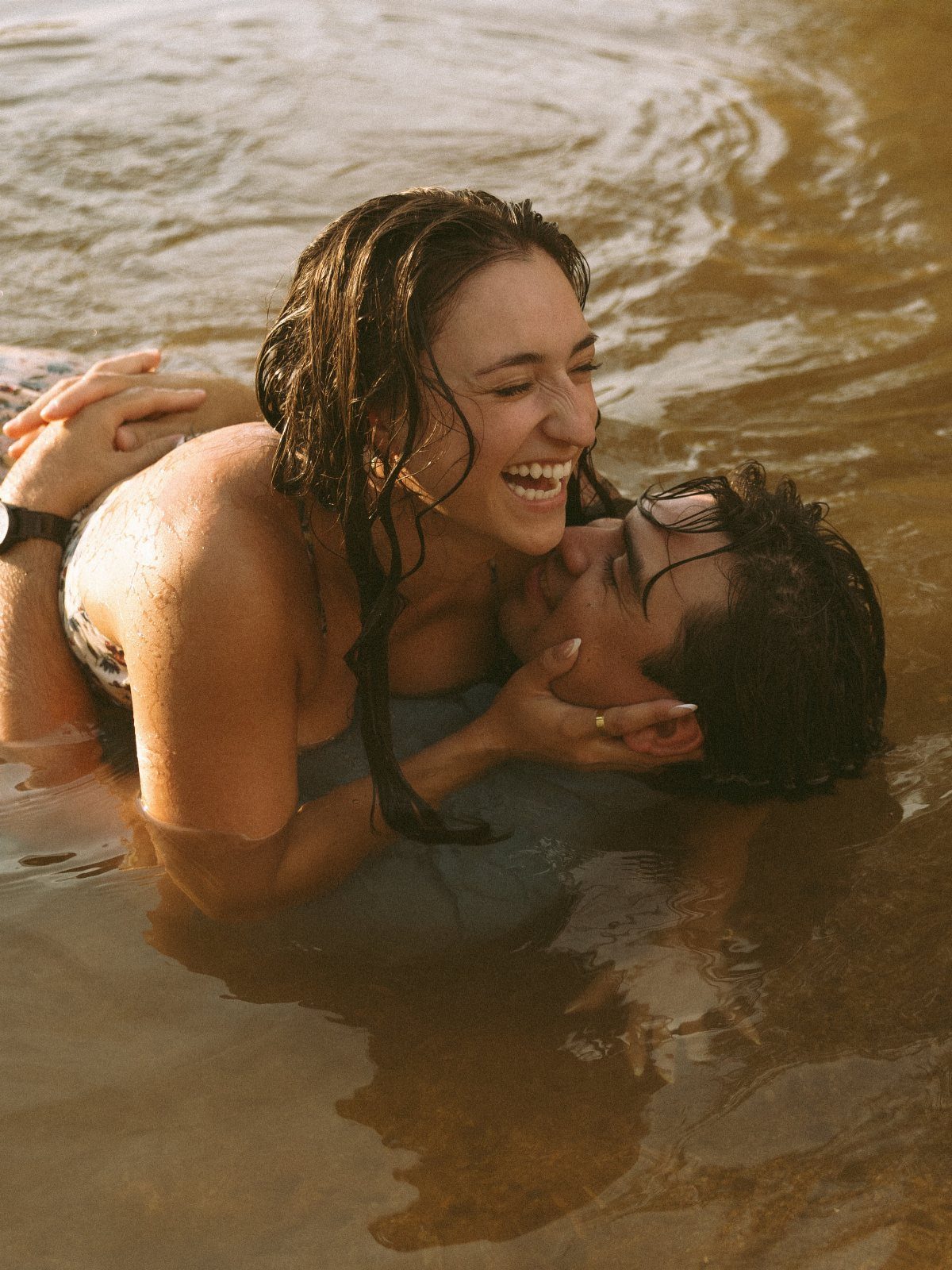 Smiling couple lying together in shallow beach water during a golden hour photoshoot.