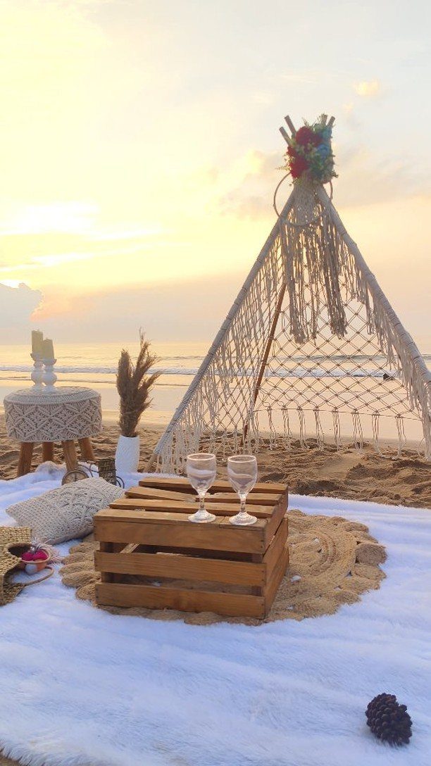 Romantic beach picnic setup with a macrame teepee and wine glasses at sunset