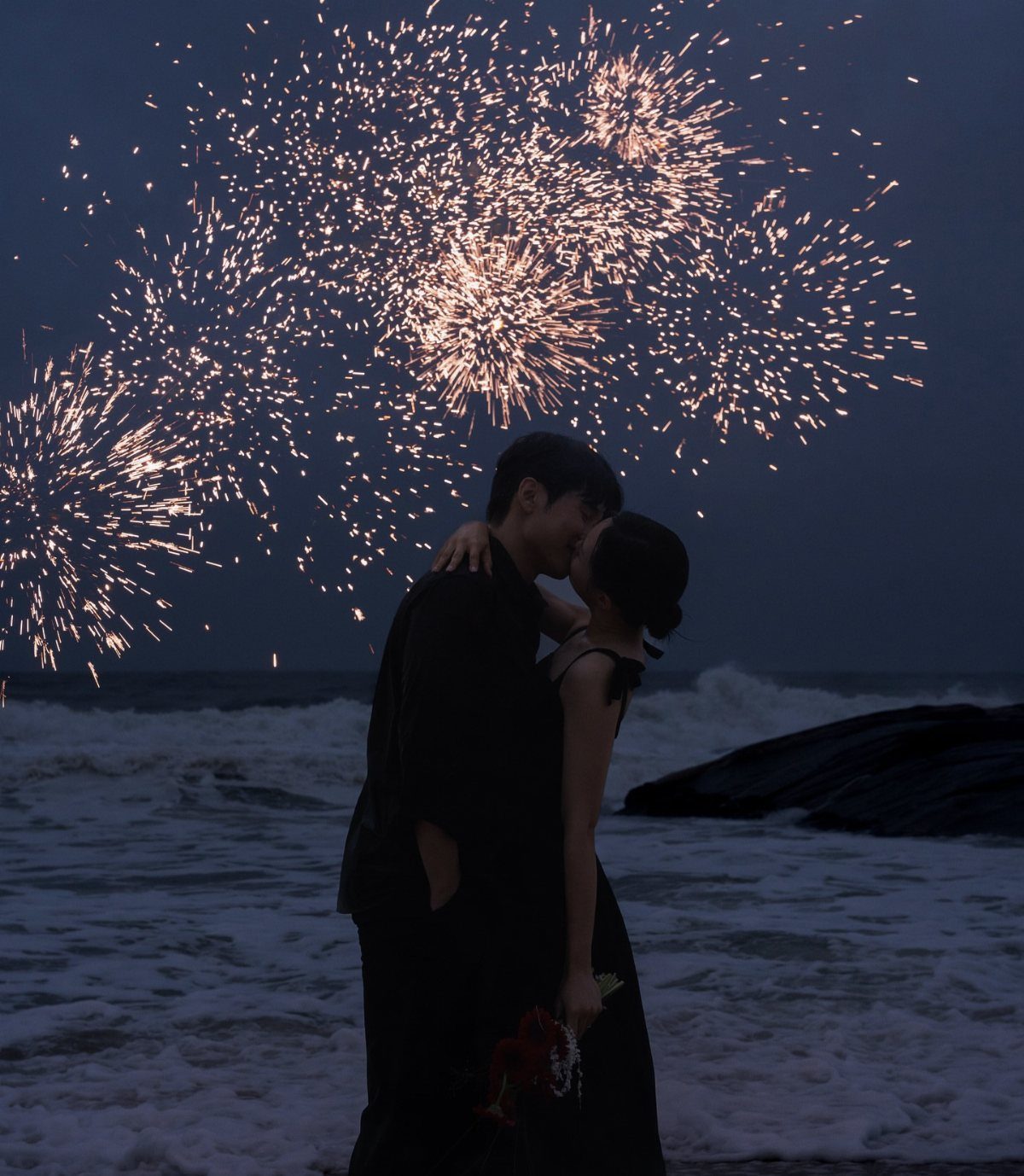 Couple kissing on a beach at night under exploding gold fireworks and waves.