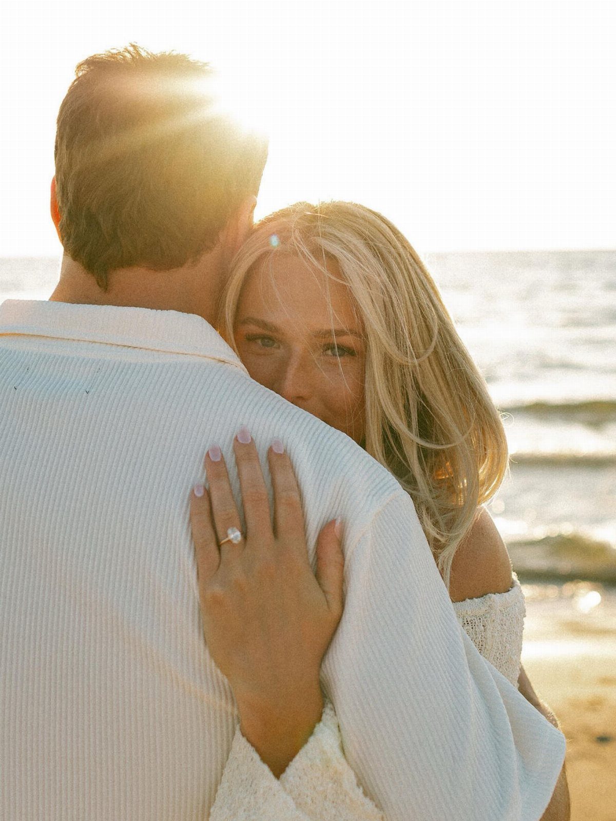 Blonde woman wearing an engagement ring hugging a man on a sunny beach