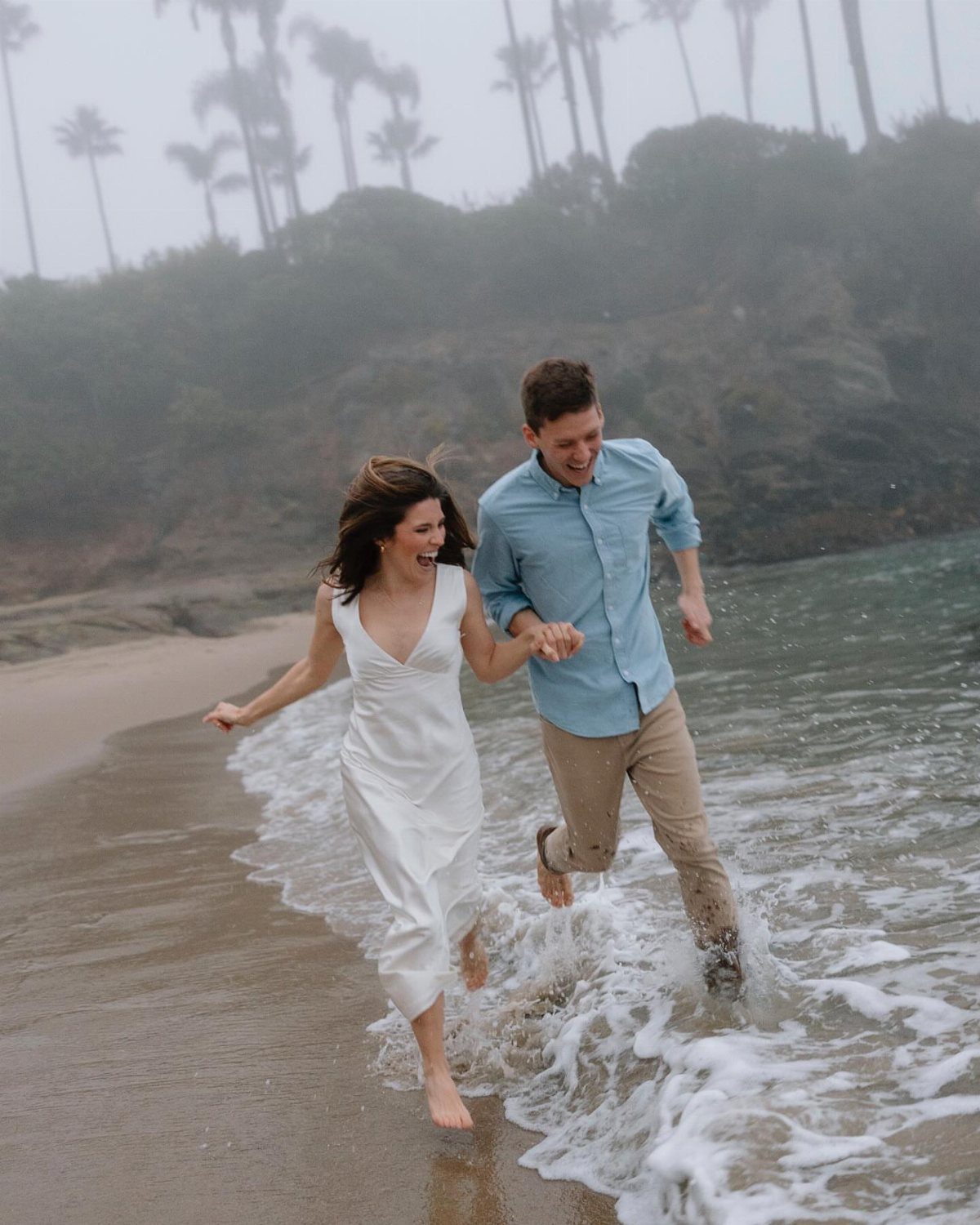 Couple running through ocean waves on a beach, woman in white flowy dress.