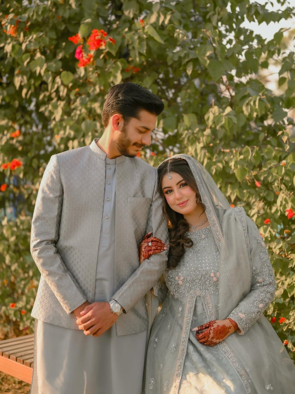 Couple in grey traditional South Asian attire posing before green garden foliage.