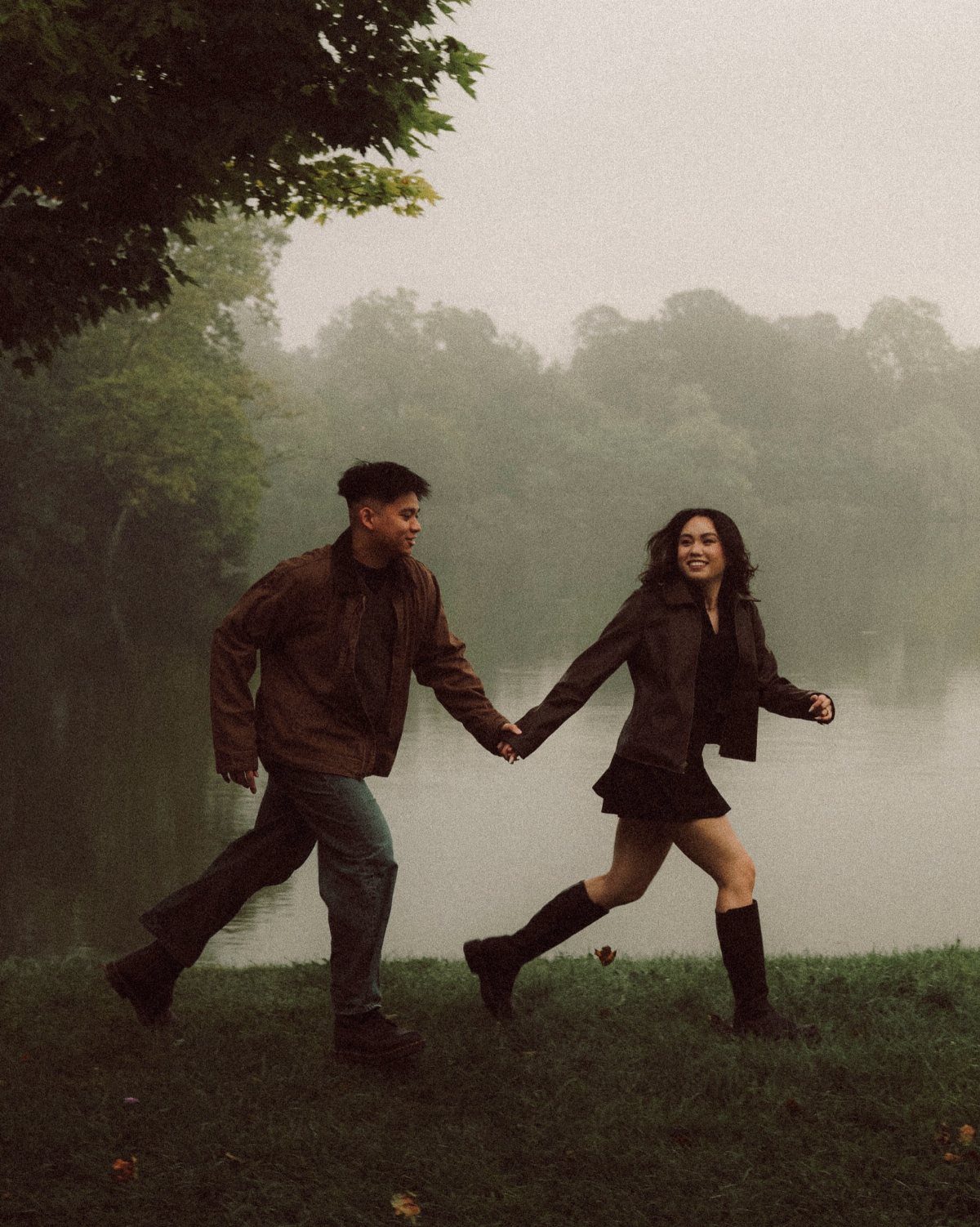 Couple walking by a lake, woman wearing a brown jacket over a black dress.