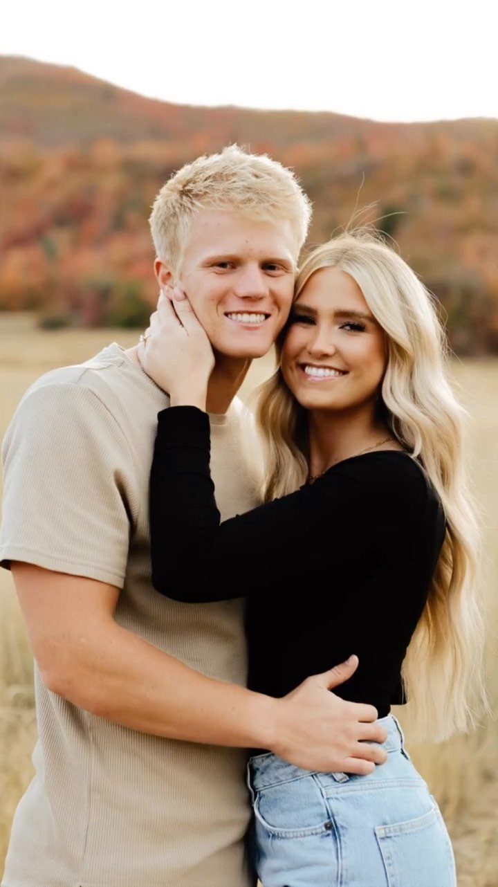 Smiling blonde couple posing in a field with autumn hills in the background
