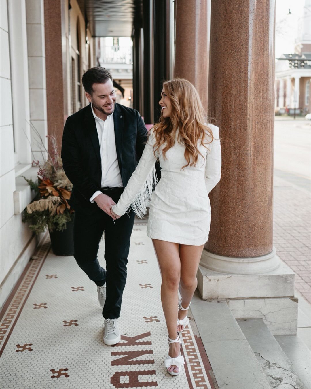 Couple walking hand-in-hand, man in blazer and sneakers, woman in short white dress.