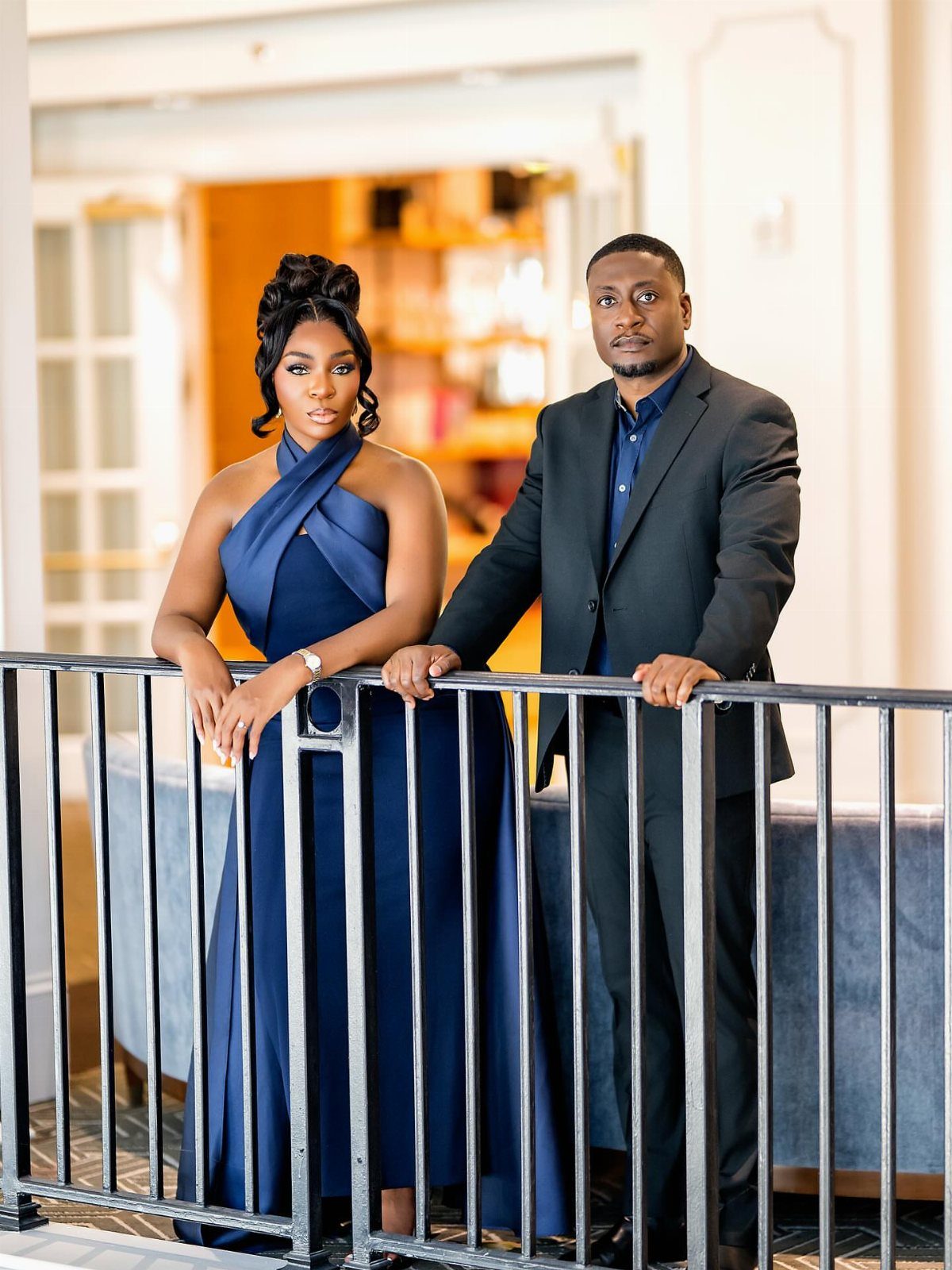 Couple posing in navy blue formal attire behind a black metal railing indoors