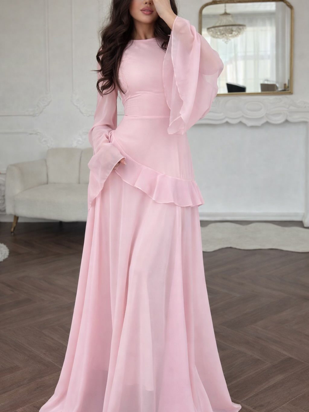 Woman in a long, flowy light pink dress with bell sleeves in a room.