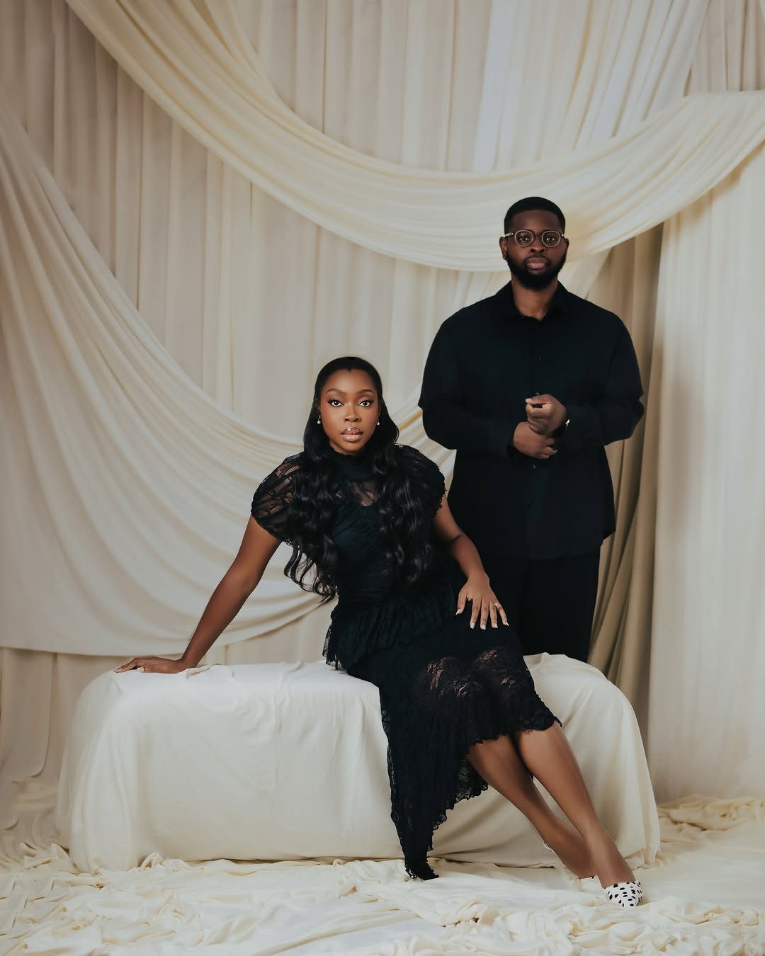 Couple posing in black formal attire against a draped cream fabric background.