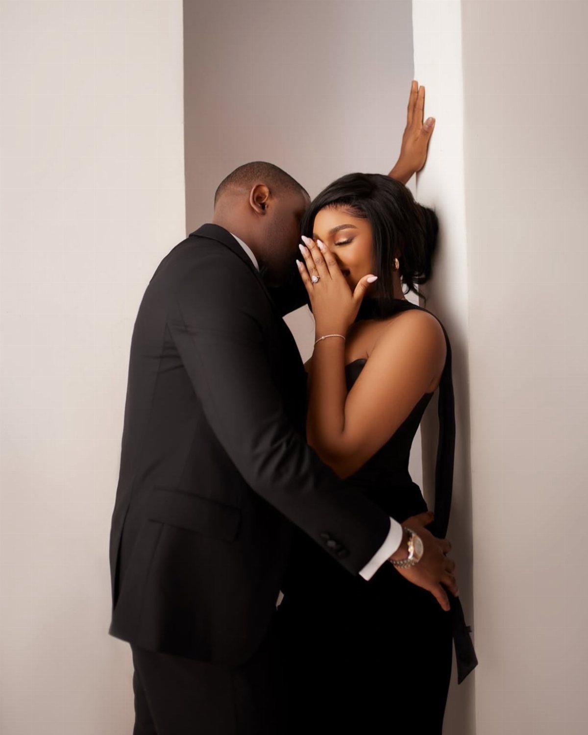 Couple in formal black attire posing closely against a plain white wall.