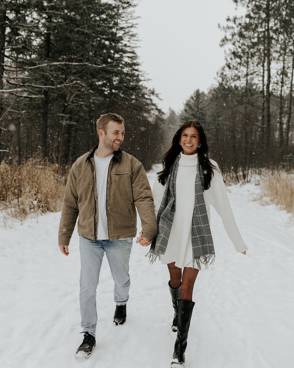 Couple walking in snow wearing winter layers including a tan jacket and plaid scarf.