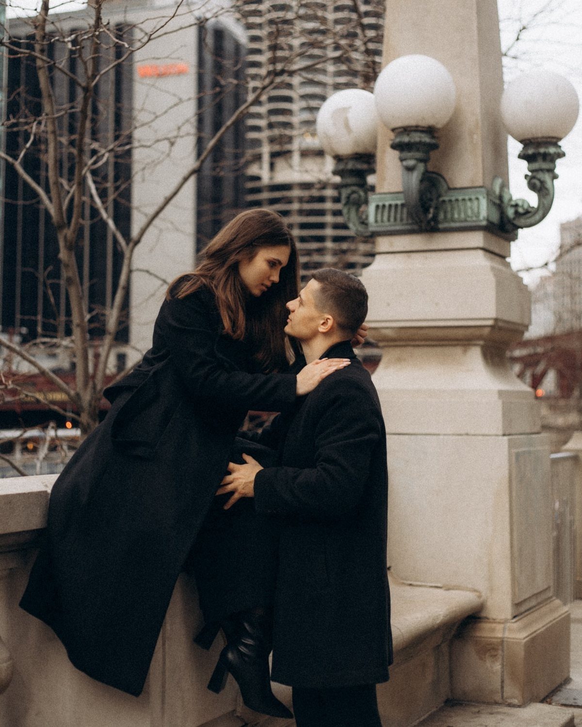 Couple in matching black coats posing romantically by a stone city lamp post.