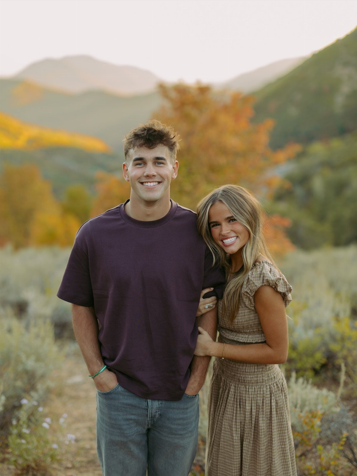 Smiling couple in autumn attire posing against a backdrop of fall foliage and mountains.