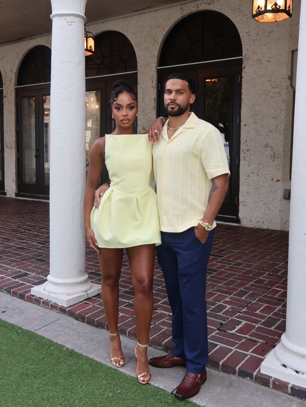 Couple posing in matching pale yellow outfits standing before a brick building.