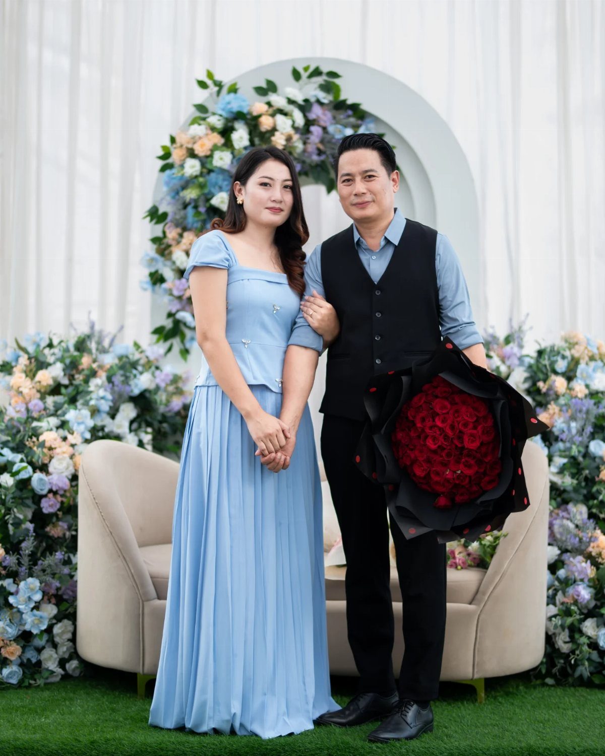 Couple posing in formal attire with a red rose bouquet and floral backdrop.