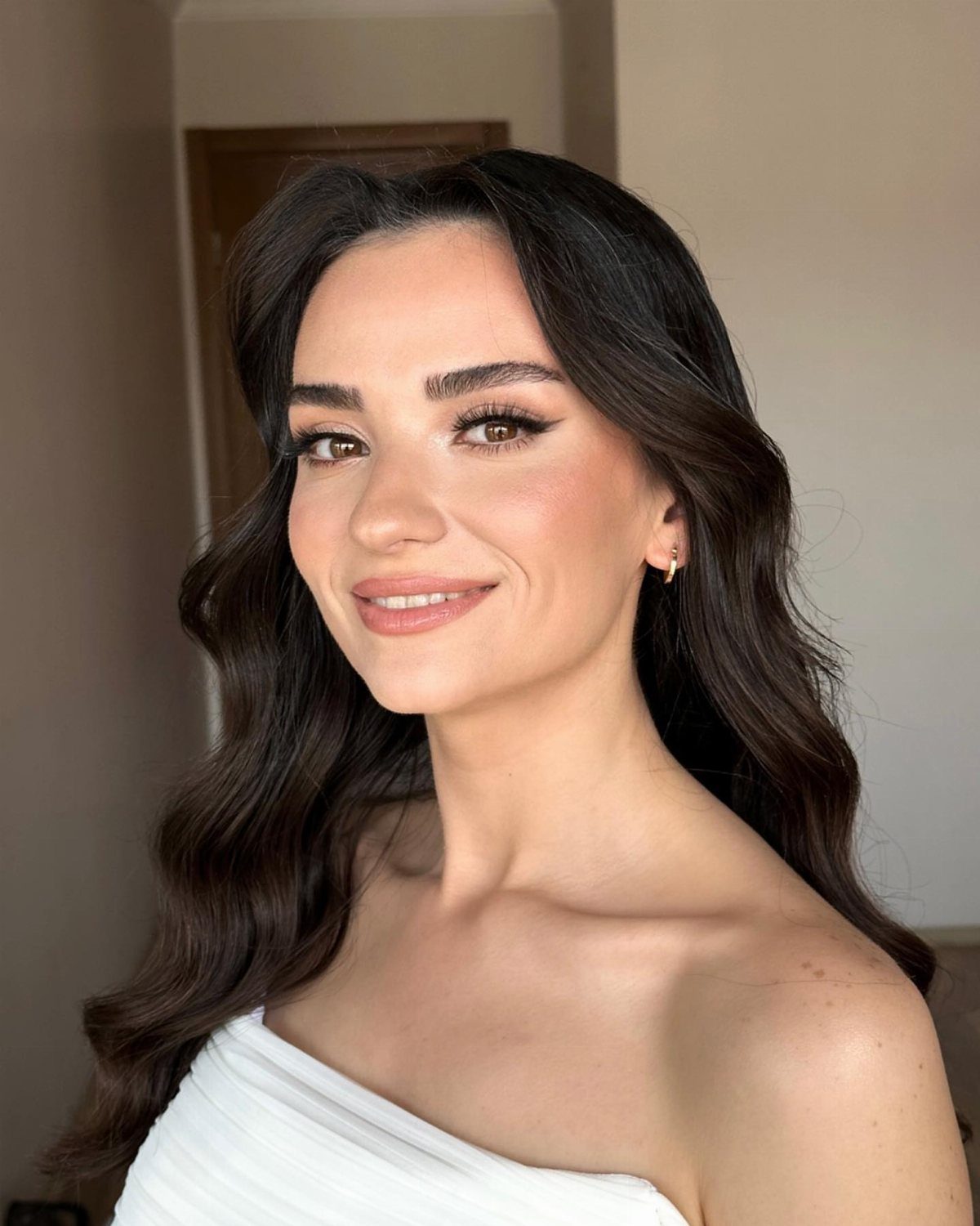 Woman with wavy dark hair and professional makeup wearing a white dress.