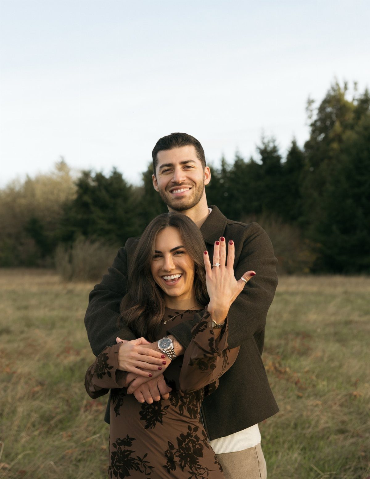 Smiling couple in brown outfits posing in a field, woman showing engagement ring