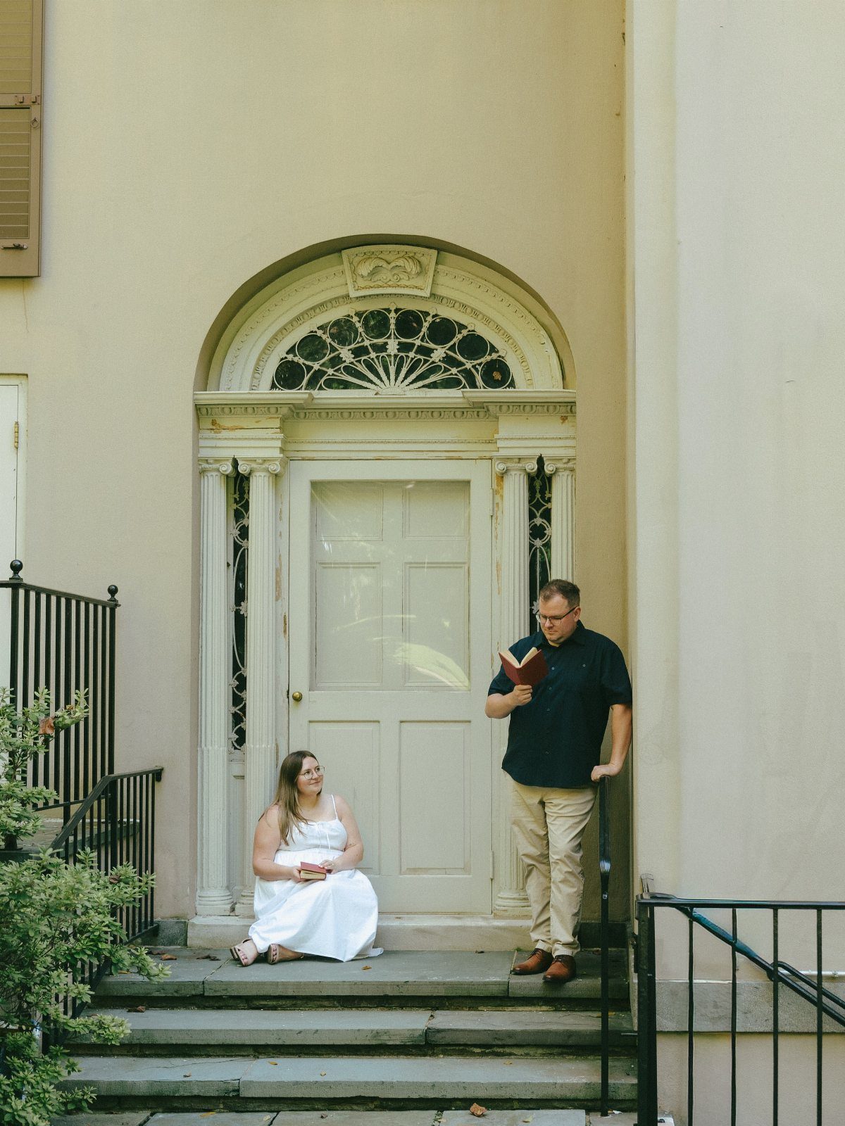 Couple posing outside a white arched doorway, woman in white dress, man reading book.