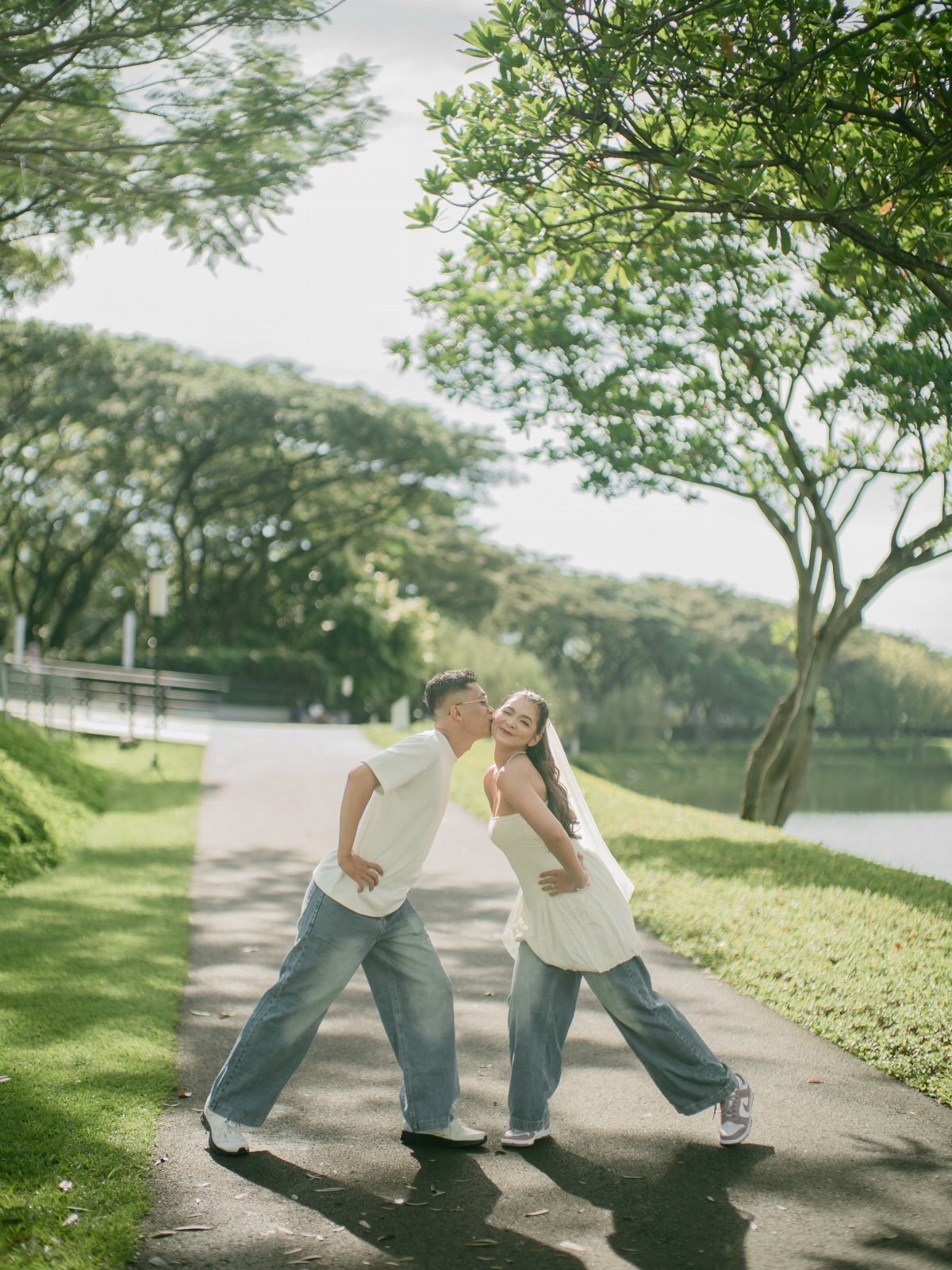 Couple in white shirts and wide-leg jeans posing on a sunny park path.